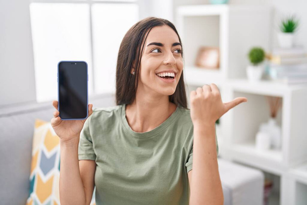młoda brunetka kobieta trzyma smartfon pokazując pusty ekran wskazując kciuk do góry na stronie uśmiechnięty szczęśliwy z otwartymi ustami  - Zdjęcie, obraz