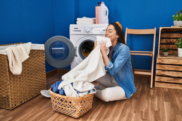 Jonge mooie Spaanse vrouw glimlachend vol vertrouwen ruikend schone kleren in de wasruimte - Foto, afbeelding