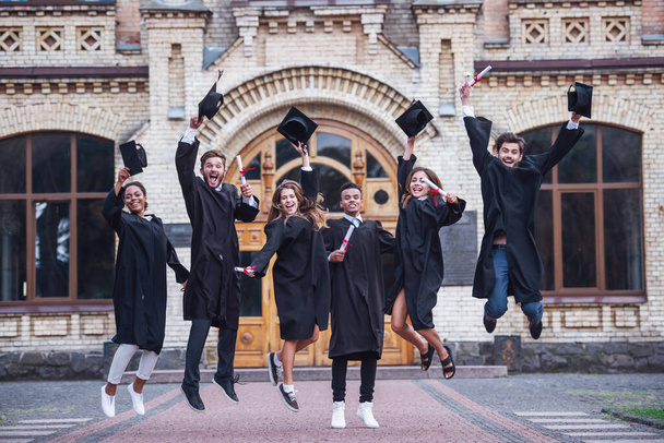 Succesvol afgestudeerden in academische jurken zijn bezit van diploma's, camera kijken en glimlachen terwijl het springen voor de foto buitenshuis - Foto, afbeelding