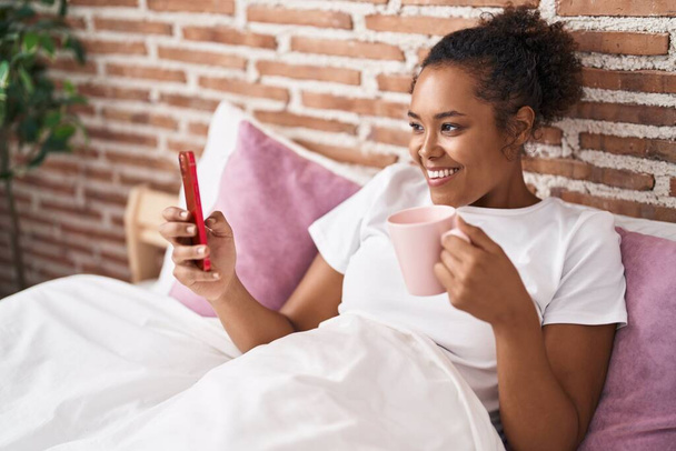 Αφροαμερικανή γυναίκα που χρησιμοποιεί smartphone πίνοντας καφέ στο υπνοδωμάτιο - Φωτογραφία, εικόνα