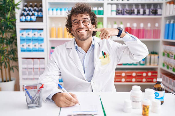Latino nuori mies työskentelee apteekissa apteekki apteekki hymyillen iloinen osoittaa ja osoittaa sormilla hampaat ja suu. hammasterveyden käsite.  - Valokuva, kuva