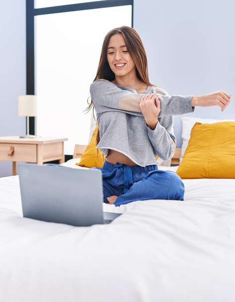 Young beautiful hispanic woman doing online stretching exercise sitting on bed at bedroom - Fotoğraf, Görsel