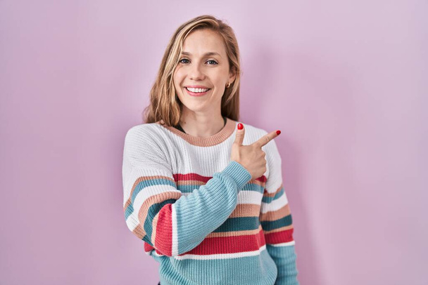 Young blonde woman standing over pink background cheerful with a smile on face pointing with hand and finger up to the side with happy and natural expression  - Foto, imagen