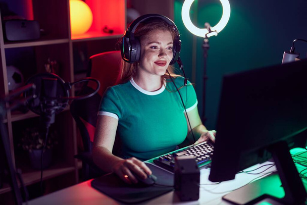 ゲームルームでコンピュータを使用してビデオゲームをプレイする若い赤毛女性ストリーマー - 写真・画像