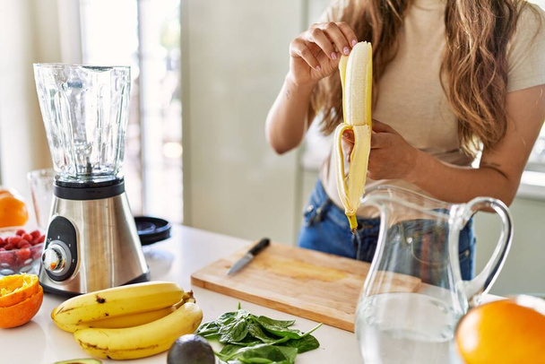 Młoda piękna Latynoska przygotowuje koktajl warzywny z mikserem obierając banana w kuchni - Zdjęcie, obraz