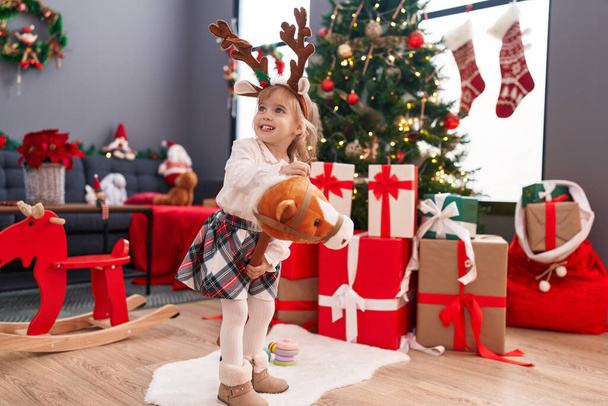 Αξιολάτρευτο ξανθό κορίτσι παίζει με το παιχνίδι άλογο στέκεται από το χριστουγεννιάτικο δέντρο στο σπίτι - Φωτογραφία, εικόνα