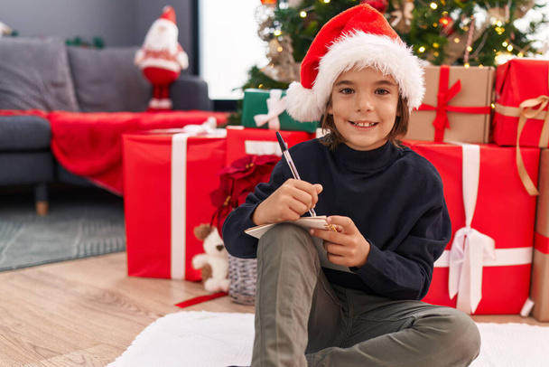 Αξιολάτρευτο ισπανικό αγόρι που γράφει γράμμα του Άη Βασίλη καθισμένο δίπλα στο χριστουγεννιάτικο δέντρο στο σπίτι. - Φωτογραφία, εικόνα
