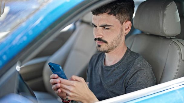 Young hispanic man using smartphone sitting on car at street - Zdjęcie, obraz