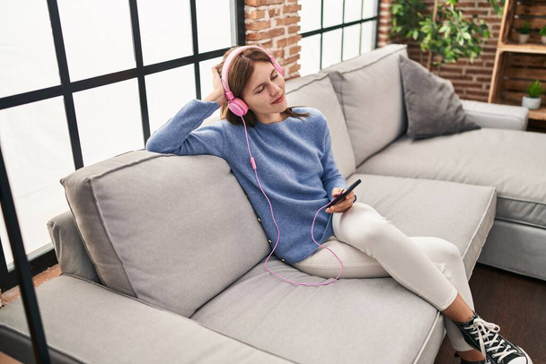 Giovane donna bionda che ascolta musica seduta sul divano di casa - Foto, immagini