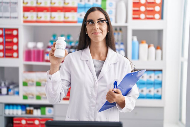 Молода брюнетка, яка працює в аптеці, тримає таблетки посміхаючись дивитися в бік і дивитися вниз думаючи.  - Фото, зображення