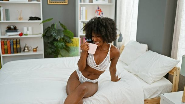 Afro-Amerikalı kadın, yatakta oturmuş kahve içiyor ve yatak odasında seksi görünüyor. - Fotoğraf, Görsel