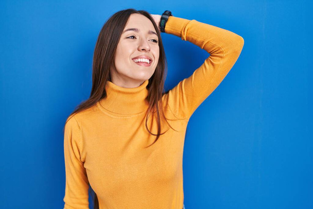 Young brunette woman standing over blue background smiling confident touching hair with hand up gesture, posing attractive and fashionable  - Photo, Image