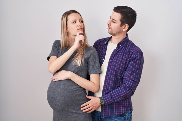 Junges Paar erwartet ein Baby, das vor weißem Hintergrund steht, die Hand am Kinn, über Fragen nachdenkend, nachdenklich. lächelndes und nachdenkliches Gesicht. Zweifelhaftes Konzept.  - Foto, Bild