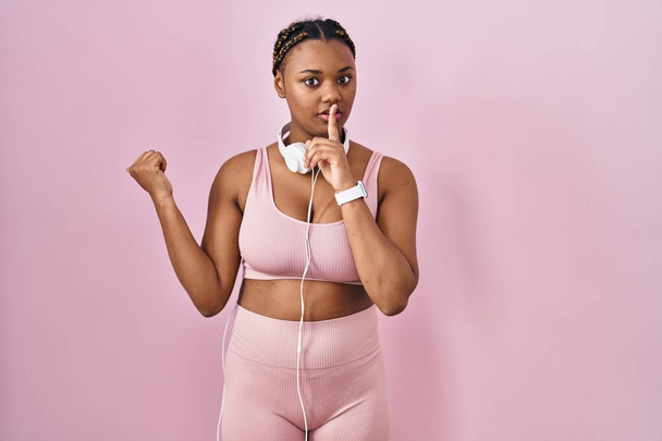 African american woman with braids wearing sportswear and headphones asking to be quiet with finger on lips pointing with hand to the side. silence and secret concept.  - Foto, Bild