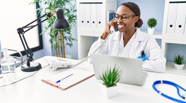 Africano americano mulher médico usando laptop falando por telefone na clínica - Foto, Imagem
