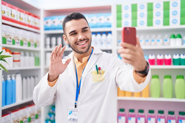 Fiatal spanyol férfi dolgozik gyógyszertár gyógyszertár videohívás okostelefon néz pozitív és boldog álló és mosolygó magabiztos mosollyal mutatja fogak  - Fotó, kép