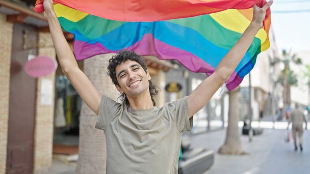 Молодий іспаномовний чоловік посміхається впевнено тримає веселковий прапор на вулиці - Фото, зображення