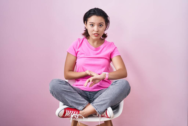 Ισπανίδα νεαρή γυναίκα κάθεται στην καρέκλα πάνω από ροζ φόντο σε βιασύνη δείχνει να παρακολουθήσουν το χρόνο, ανυπομονησία, αναστατωμένος και θυμωμένος για καθυστέρηση προθεσμία  - Φωτογραφία, εικόνα