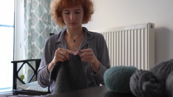 teenage girl with curly red hair, sitting at home in pajamas on chair, knits product from gray woolen thread. - Footage, Video