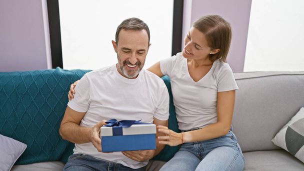 Father's loving surprise gift leaves birthday girl, his daughter, smiling and happily surprised on their casual, relaxing living room sofa at home. - Photo, Image