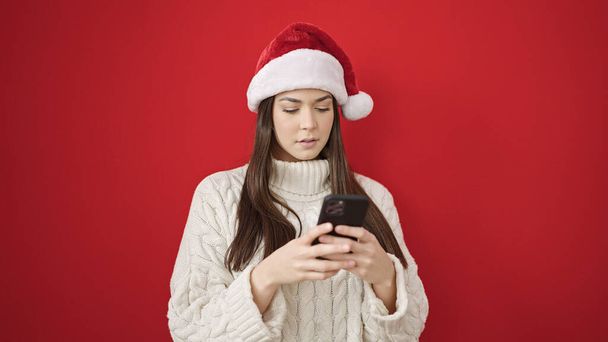 Młoda, piękna Latynoska kobieta w świątecznym kapeluszu za pomocą smartfona na izolowanym czerwonym tle - Zdjęcie, obraz