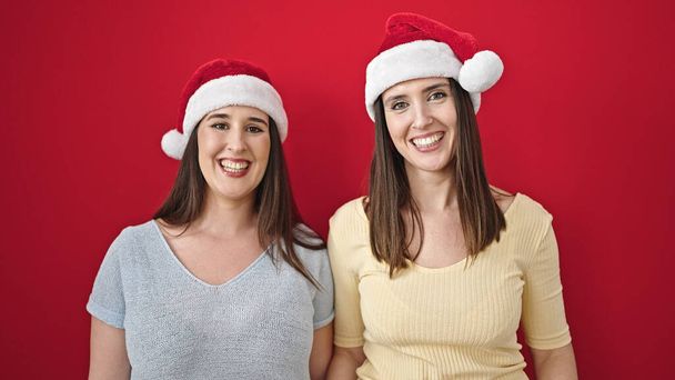 Dos mujeres sonriendo confiadas usando sombrero de Navidad sobre fondo rojo aislado - Foto, Imagen