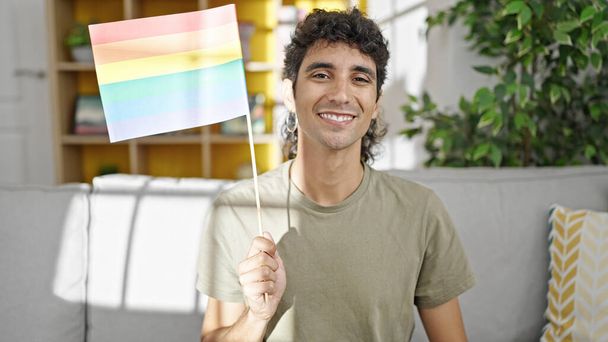 Joven hombre hispano sosteniendo bandera de arco iris sentado en el sofá en casa - Foto, Imagen