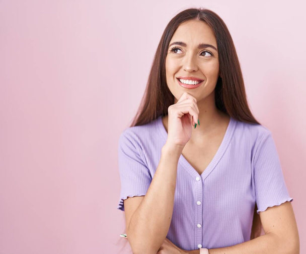 Jonge Latijns-Amerikaanse vrouw met lang haar staande over roze achtergrond met de hand op kin denken over vraag, pensive expressie. glimlachend en attent gezicht. twijfelconcept.  - Foto, afbeelding