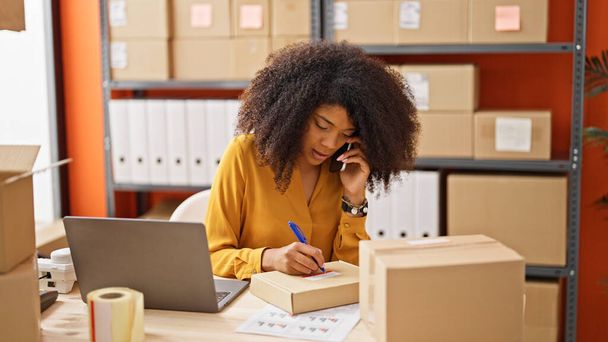 Αφρο-αμερικανίδα υπάλληλος ηλεκτρονικού εμπορίου μιλώντας στο smartphone γράφοντας στο πακέτο στο γραφείο - Φωτογραφία, εικόνα