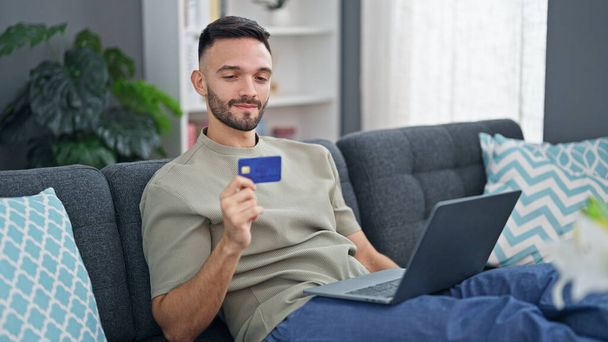 Молодой латиноамериканец с ноутбуком и кредитной картой сидит дома на диване - Фото, изображение