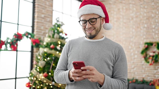 Homem hispânico usando chapéu de Natal usando smartphone em casa - Foto, Imagem