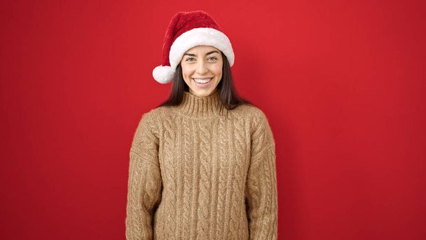 Junge schöne hispanische Frau lächelt selbstbewusst mit Weihnachtsmütze über isoliertem rotem Hintergrund - Foto, Bild