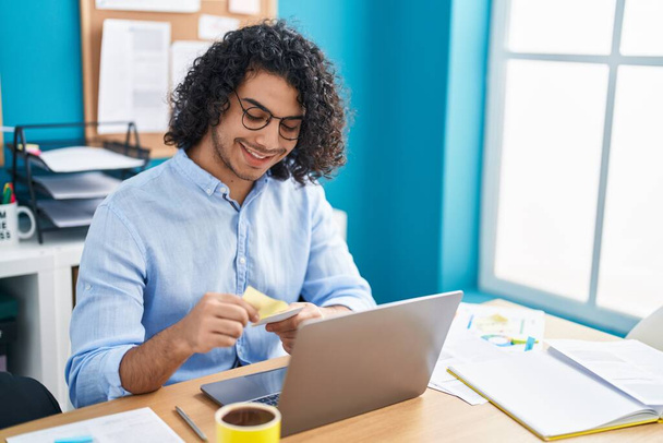 Young latin man business worker using laptop holding reminder paper at office - Photo, Image