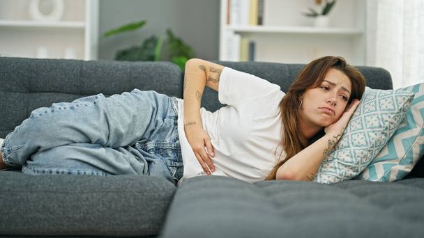 Νεαρή όμορφη ισπανόφωνη γυναίκα που υποφέρει για εμμηνορροϊκό πόνο ξαπλωμένη στον καναπέ στο σπίτι - Φωτογραφία, εικόνα