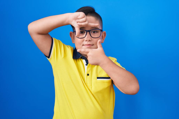 Young hispanic kid standing over blue background smiling making frame with hands and fingers with happy face. creativity and photography concept.  - Foto, afbeelding
