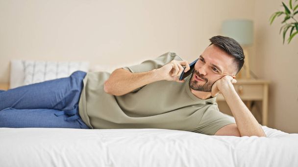 Fiatal spanyol férfi beszél okostelefonon fekszik az ágyon a hálószobában - Fotó, kép