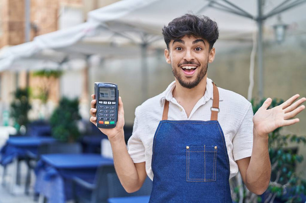 Araber mit Bart trägt Kellnerschürze auf Restaurantterrasse und hält Datafon in der Hand, um Leistung mit glücklichem Lächeln und Siegerausdruck mit erhobener Hand zu feiern  - Foto, Bild