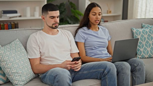 Hermosa pareja enamorada, sentados juntos en el sofá de la sala de estar en casa, absortos en la tecnología. hombre y mujer usando su computadora portátil y teléfono inteligente, trabajando y mensajes de texto en línea. - Foto, imagen