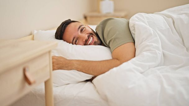 Fiatal spanyol férfi mosolyogva magabiztos feküdt az ágyon a hálószobában - Fotó, kép
