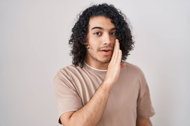 Homme hispanique avec les cheveux bouclés debout sur fond blanc main sur bouche racontant rumeur secrète, chuchotant conversation malicieuse  - Photo, image
