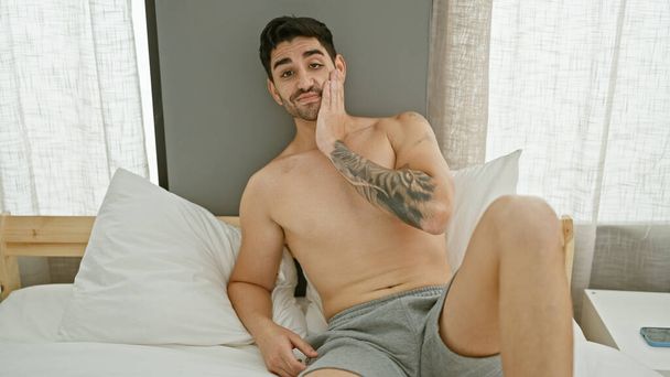 Joven hombre hispano acostado en la cama con expresión relajada en el dormitorio - Foto, Imagen