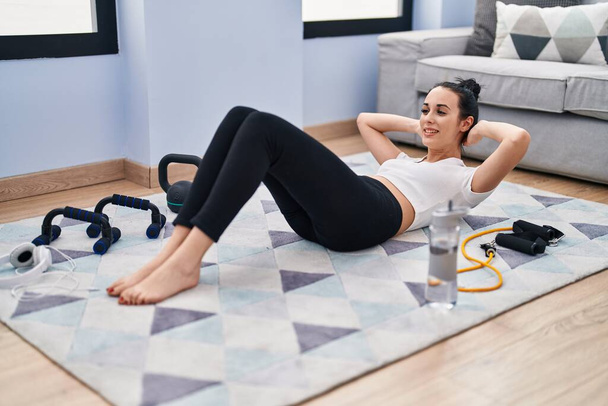 Молодая белая женщина улыбается уверенно тренировки пресса упражнения на дому - Фото, изображение