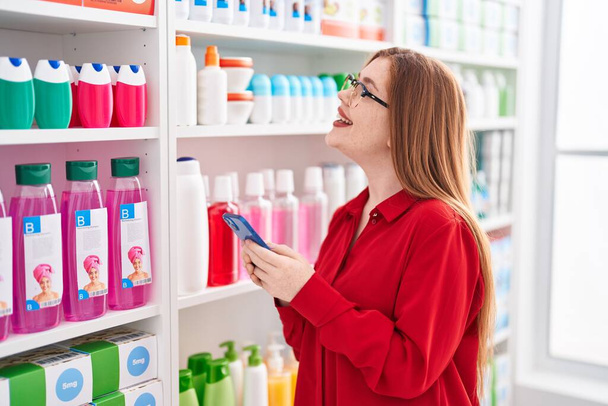 Jeune rousse femme client souriant confiant en utilisant smartphone à la pharmacie - Photo, image