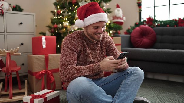 Kafkasyalı genç adam akıllı telefon kullanıyor. Evdeki Noel ağacının yanında yerde oturuyor. - Fotoğraf, Görsel
