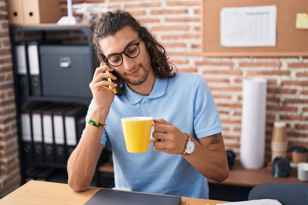 Молодой латиноамериканец-бизнесмен разговаривает по смартфону, пьет кофе в офисе - Фото, изображение