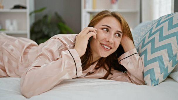 Νεαρή γυναίκα μιλάει στο smartphone ξαπλωμένη στο κρεβάτι στο υπνοδωμάτιο - Φωτογραφία, εικόνα