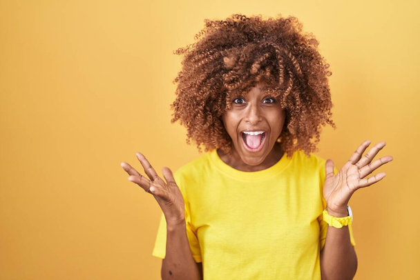 Jeune femme hispanique avec les cheveux bouclés debout sur fond jaune célébrant fou et étonné pour le succès avec les bras levés et les yeux ouverts hurlant excités. concept gagnant  - Photo, image