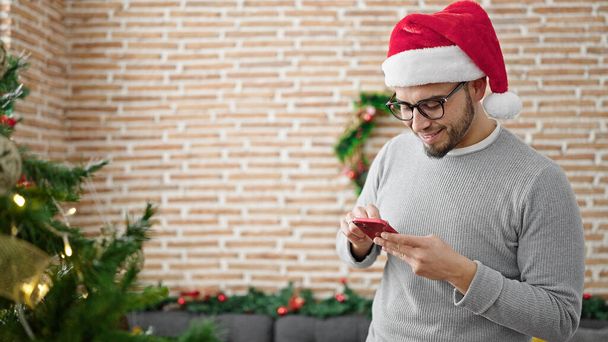 Uomo ispanico che indossa il cappello di Natale utilizzando smartphone a casa - Foto, immagini