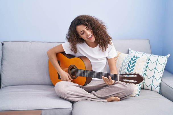 Young beautiful hispanic woman playing classical guitar sitting on sofa at home - Photo, Image