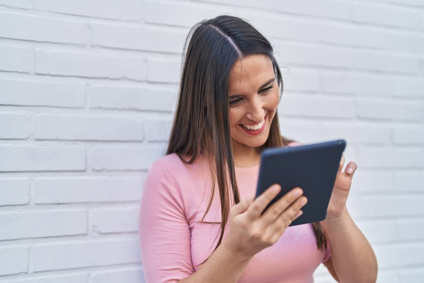 Jovem bela mulher hispânica sorrindo confiante usando touchpad sobre isolado fundo de tijolo branco - Foto, Imagem